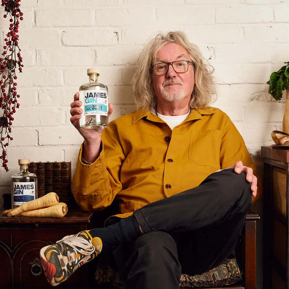 A man with gray hair and glasses in a mustard-yellow jacket and colorful sneakers sits cross-legged indoors, smiling at the camera while holding James Gin US The Six Pack Asian Parsnip & Navy Strength with a souvenir gift key chain. Another bottle is nearby.