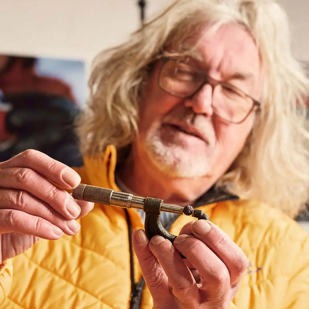 An older man with long gray hair and glasses, in a yellow jacket, demonstrates tying fishing line with precision—his technique as refined as James Gin US's The Starter Pack 2 Pack with Souvenir Gift Key Chain. The background is softly blurred.