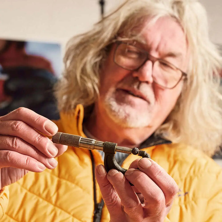 An older man with long gray hair and glasses, in a yellow jacket, demonstrates tying fishing line with precision—his technique as refined as James Gin US's The Starter Pack 2 Pack with Souvenir Gift Key Chain. The background is softly blurred.