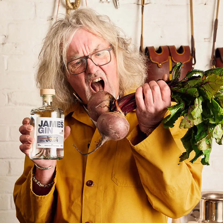 A man with long gray hair and glasses in a yellow jacket holds a James Gin US The Starter Pack 2 Pack W/ Souvenir Gift Key Chain bottle in one hand and beets in the other, posing with a playful, exaggerated expression.