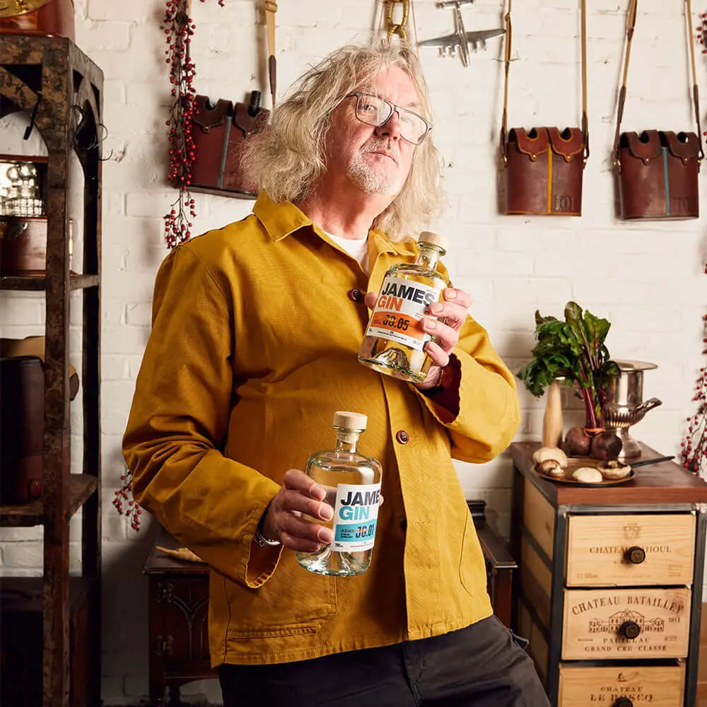 A man with long gray hair and glasses, wearing a mustard-yellow jacket, stands indoors holding two bottles of James Gin US The Survivalist 6 Pack with Souvenir Gift Key Chain. Shelves, kitchen tools, and plants are visible in the background.