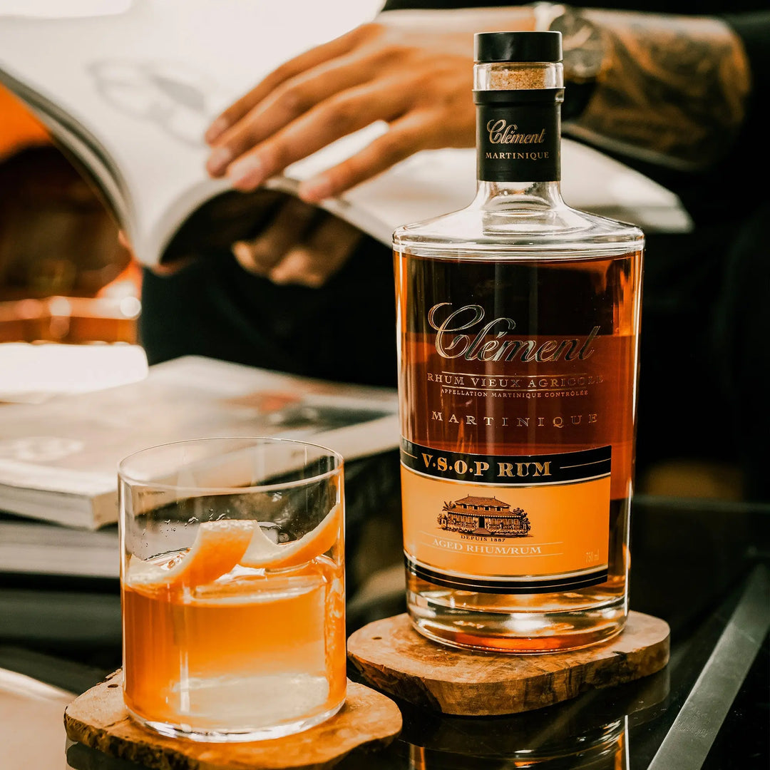 A bottle of Rhum Clément V.S.O.P. and a glass of AOC Martinique rum garnished with orange peel rest on wooden coasters, as someone reads a book in the blurred background.