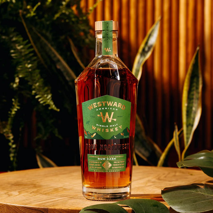 A bottle of Westward American Single Malt Rum Cask by Westward rests on a wooden surface, surrounded by green plants and warm wood tones. The label features green with gold and white text.